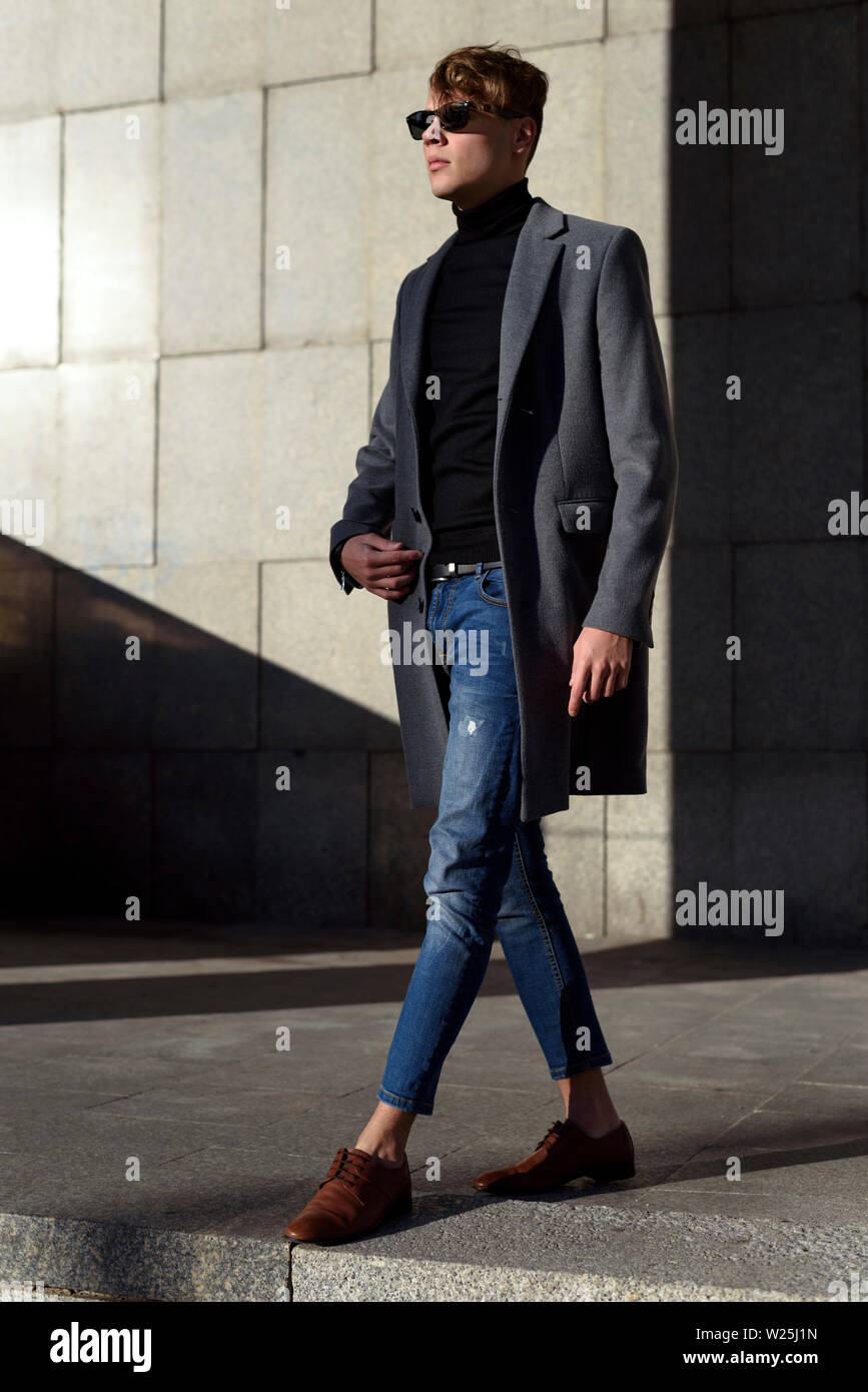 Moda joven hombre adulto en gafas de sol. Posando en el elegante chaqueta  gris, negro polo cuello, pantalones de mezclilla y brown mocasin zapatos.  Las tendencias de la moda Fotografía de stock -