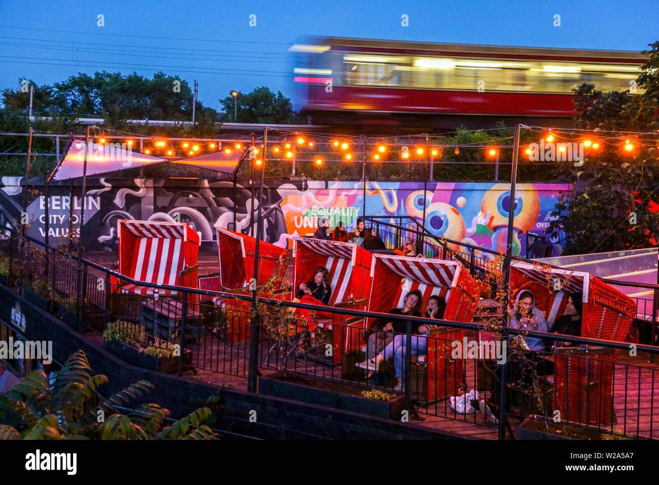 Jóvenes en el bar Else, un tren, S-Bahn va alrededor, crepúsculo, Alt Treptow, vida de la ciudad de Berlín Alemania, gente sentada en sillas de playa de mimbre Foto de stock