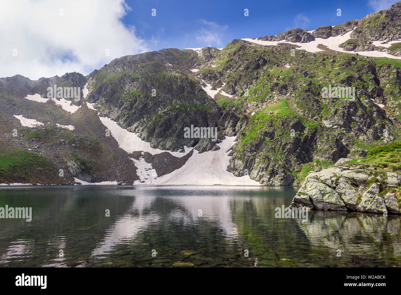 La pasto verde sobre un altiplano rocoso y picos del macizo de Rila y el reflejo en el agua limpia del Ojo del lago glacial, uno de los siete lagos de Rila Foto de stock