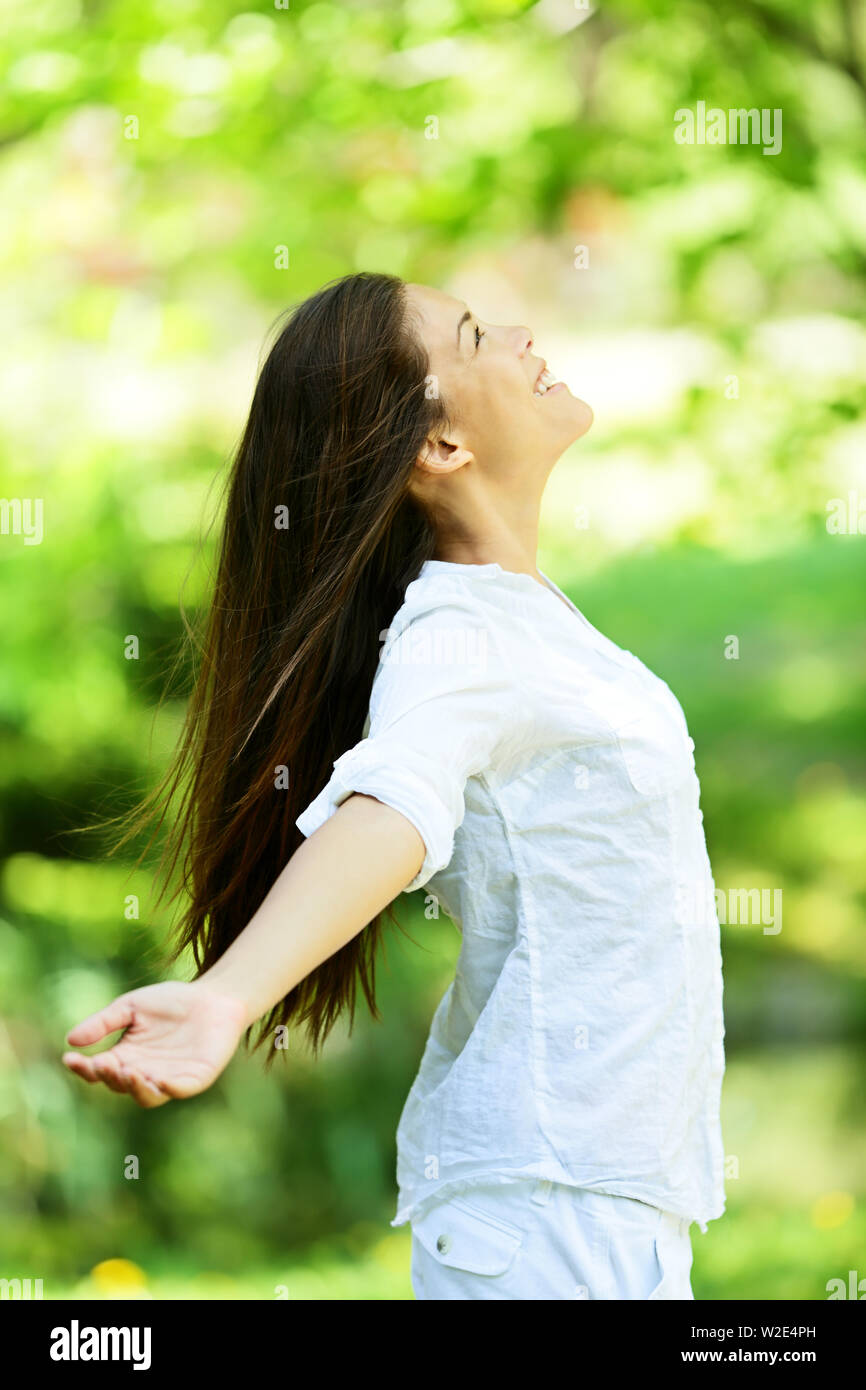 Mujer joven abrazando la llegada de la primavera, de pie en un frondoso parque, con sus brazos extendidos y su cabeza levantada hacia el cielo como exulta en la naturaleza. Concepto de diversión y libertad. Foto de stock