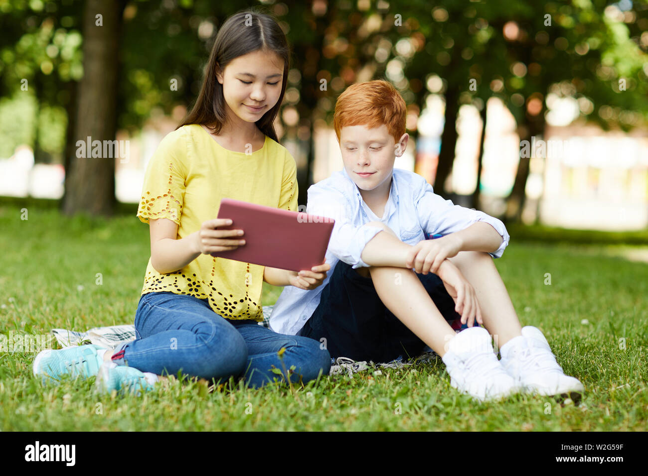 Sonriendo colegiala camiseta amarilla sentados en el césped y mostrando su proyecto sobre la tableta a su classmate Foto de stock