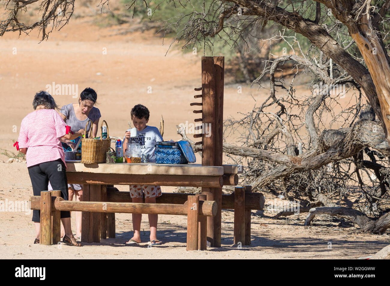 Grupo familiar en un banco de madera tabla preparando una comida en un picnic o sitio de descanso en el medio del desierto el Parque Transfronterizo Kgalagadi, Sudáfrica Foto de stock