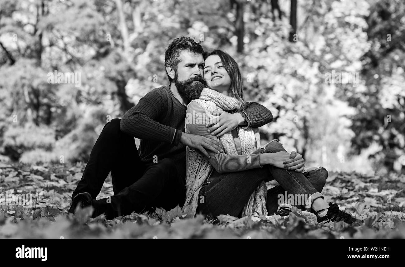 Pareja en amor con bufandas sentada sobre las hojas caídas en el parque. El  hombre y la mujer con rostros sonrientes en otoño los árboles de fondo.  Romance y soleado otoño concepto.