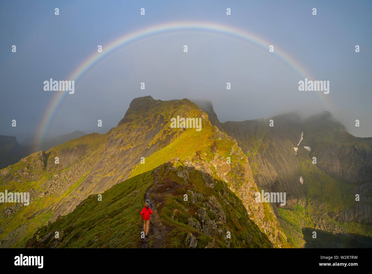 Hombre subiendo hacia un arco iris en las islas Lofoten. remoto paisaje niebla y picos de montaña Foto de stock