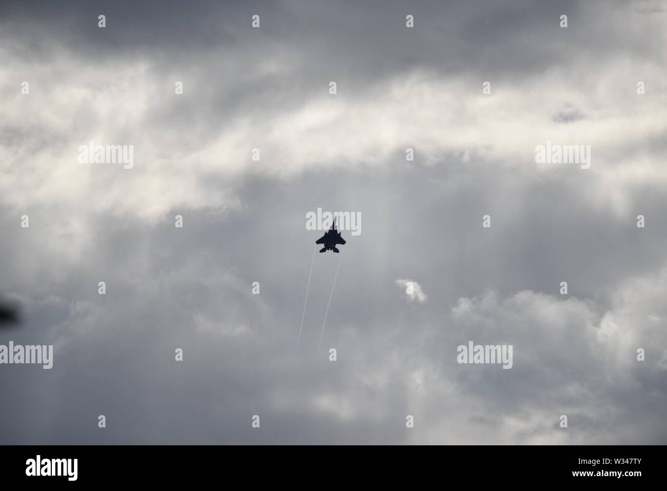 F 15 jet fighter en la distancia subiendo en un gris oscuro cielo nublado con sol casi abstracto patrones de fondo. Foto de stock