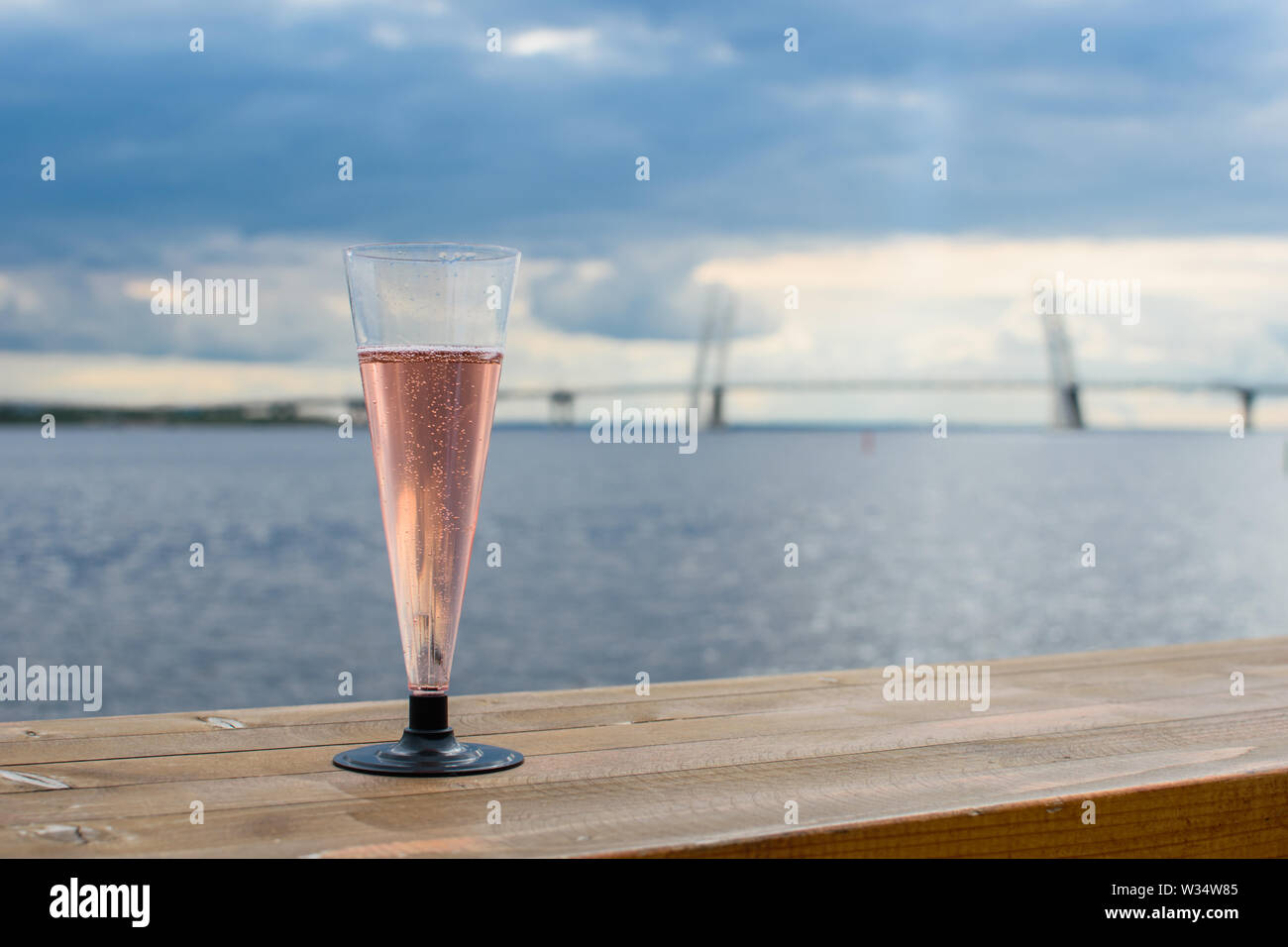 Copa de champaña de plástico sobre una superficie de madera sobre el fondo de la ciudad, el puente grande, el mar y el cielo oscuro. Foto de stock