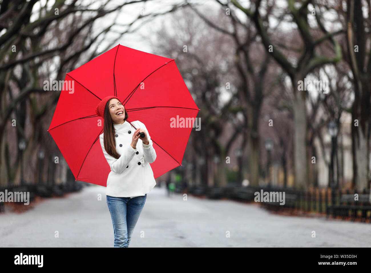 Mujer con sombrilla roja caminar en el parque en otoño. Feliz sonriente joven multirracial caminando alegre con sombrilla roja en Central Park, Manhattan, Ciudad de Nueva York, EE.UU. Foto de stock