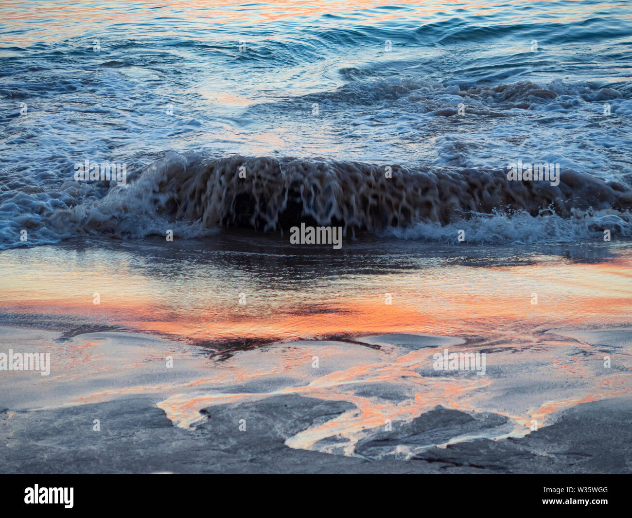 Cerca de una ola rompiendo en una playa arenosa con el reflejo del sol en el agua Foto de stock