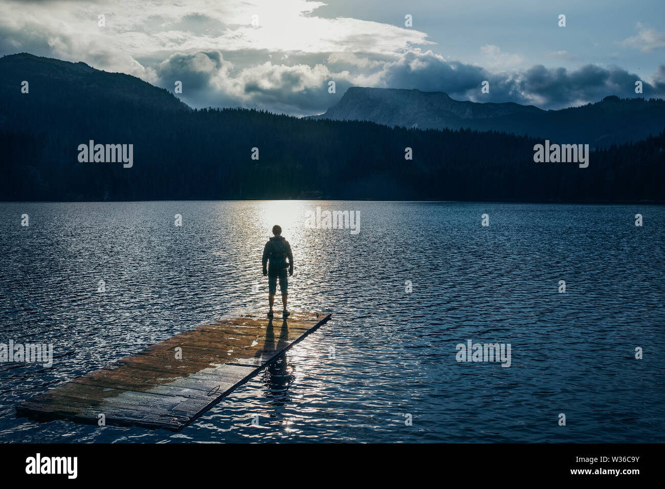 Silueta hombre de pie sobre la balsa de madera sobre el lago azul. Montañas nubladas en la espalda con la luz del atardecer en la naturaleza del paisaje Foto de stock