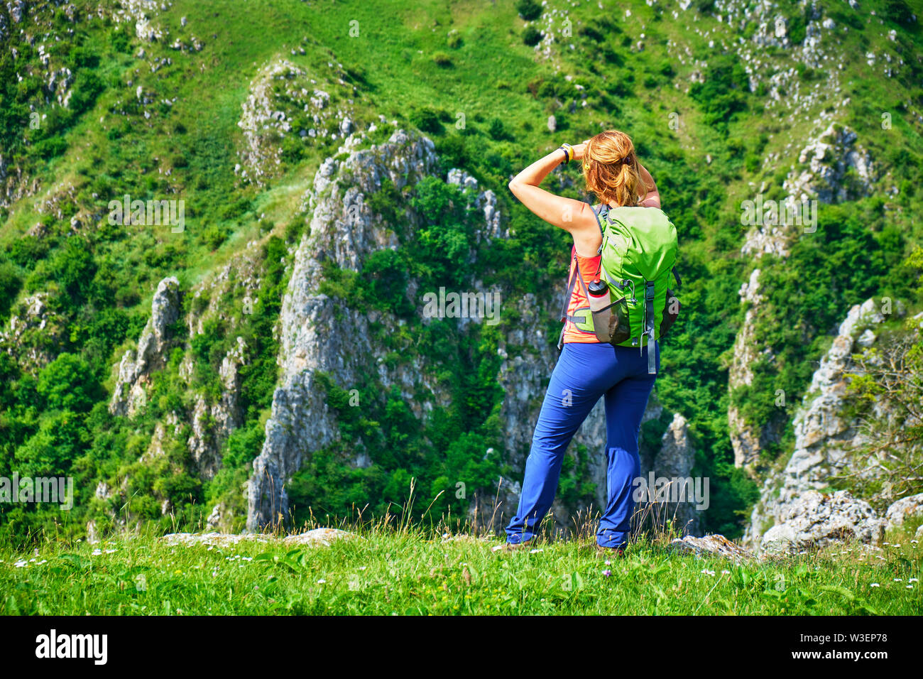 Mujer joven excursionista con una mochila verde se levanta sobre una cresta de hierba y mira hacia las enormes paredes de roca en Tureni Gorge, condado de Cluj, durante una soleada Foto de stock