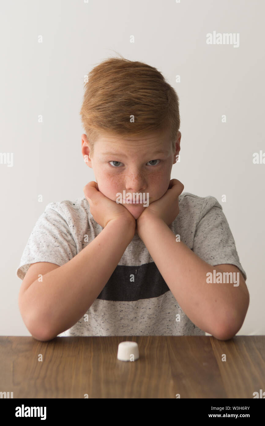 Rojo-cabeza muchacho caucásico se sienta en una mesa con un único marshmallow, intentar la prueba de malvavisco Foto de stock