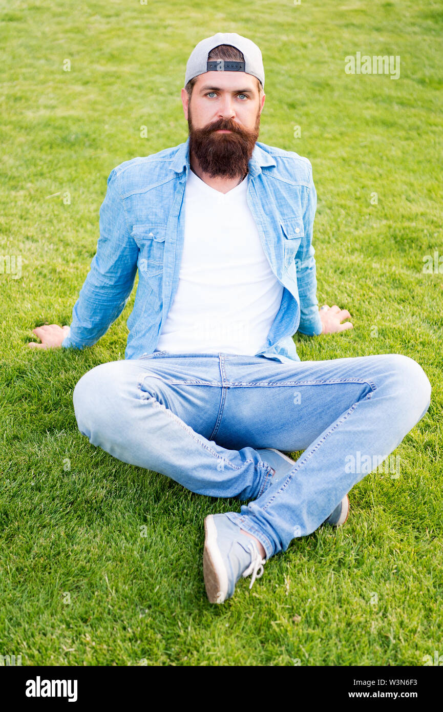 Hipster peluda. Brutal hipster sentada sobre la hierba verde. Hombre  Barbado en un moderno estilo HIPSTER en verano al aire libre. Caucasian  hipster con espesa barba cabello llevaba una gorra de béisbol