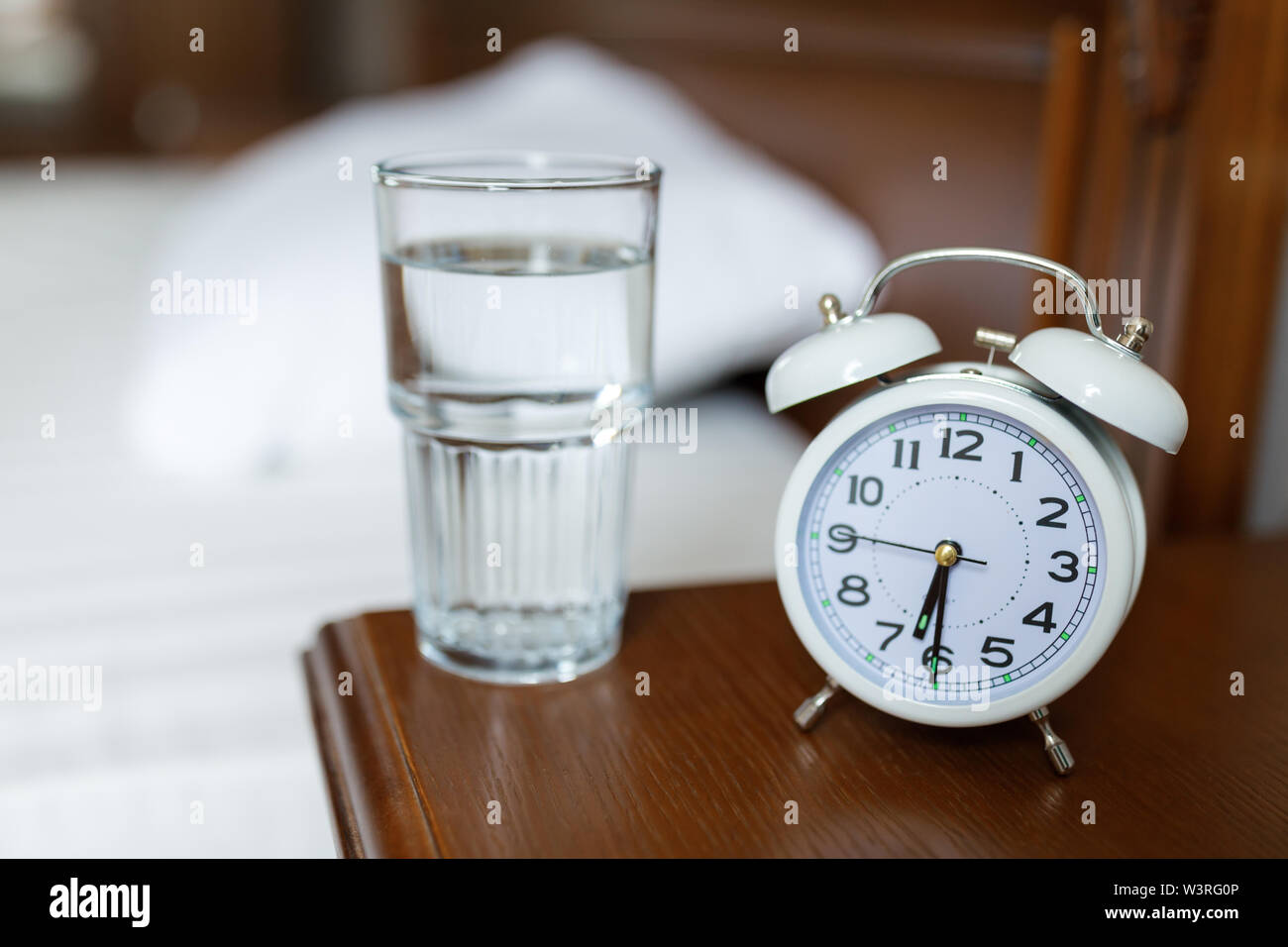 Un reloj despertador blanco y un vaso de agua en una mesita de madera. Foto de stock