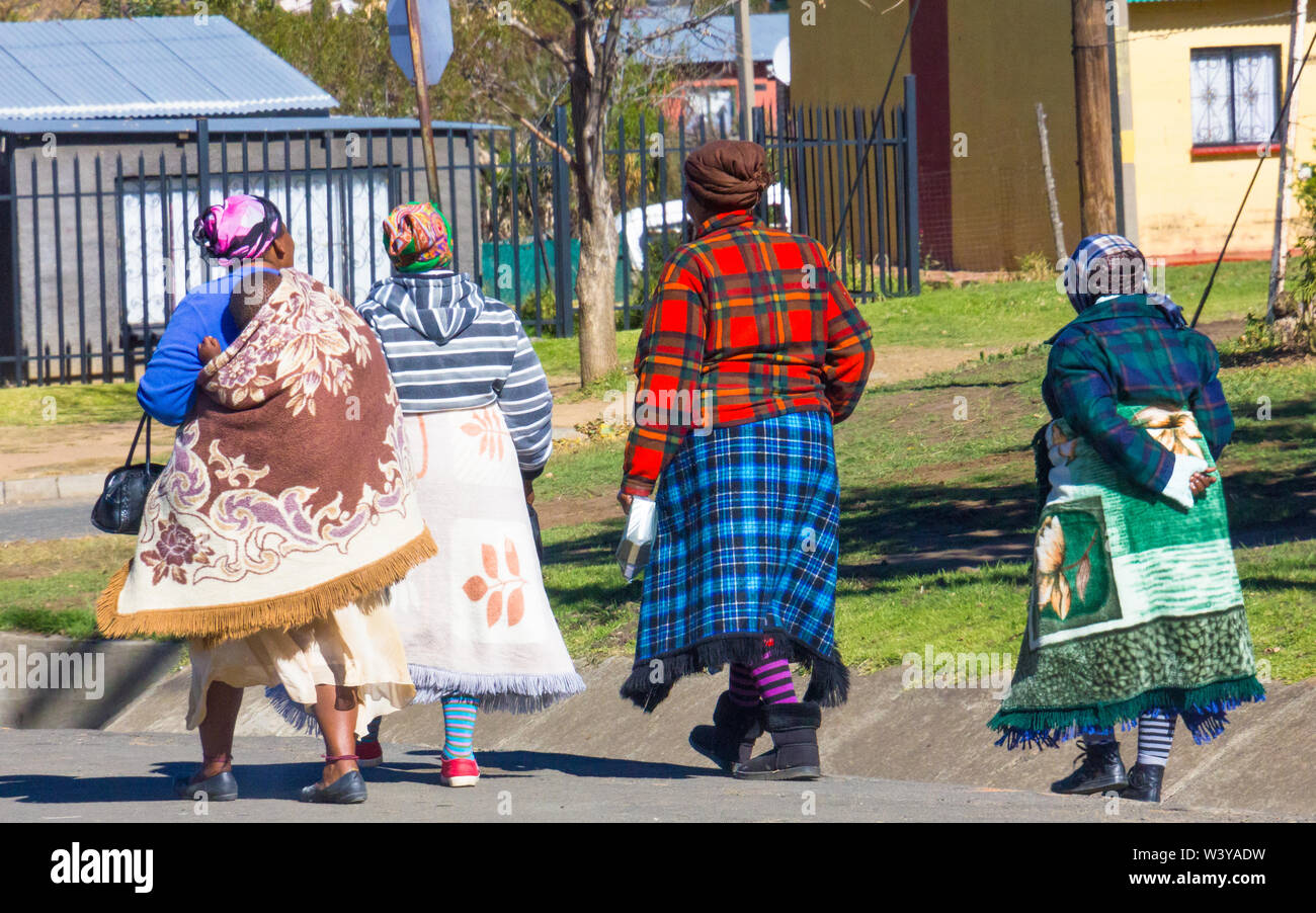 Cuatro mujeres africanas negras de origen Basotho envueltos en mantas de estilo tradicional, caminando en una calle de Clarens, Estado Libre de Orange, Sudáfrica Foto de stock