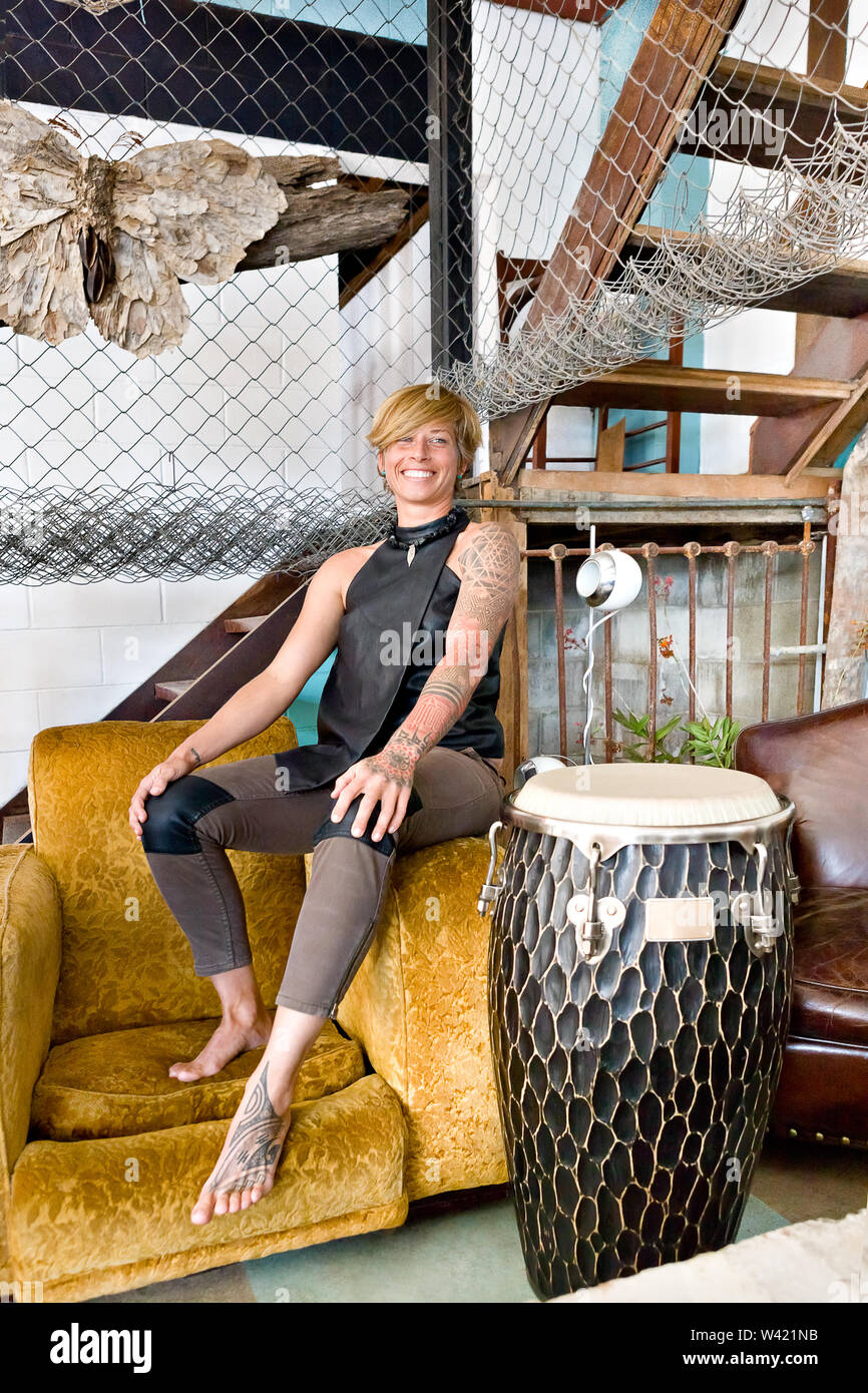Chica con una chaqueta negra y el tatuaje se sienta en un sofá de color amarillo al lado de un Bongo o tambor bajo una habitación con net y escalera, escaleras Foto de stock