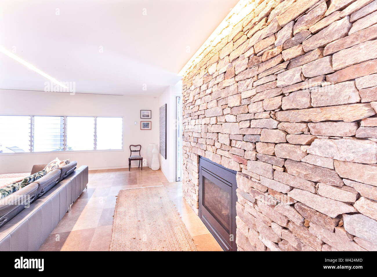 Moderna sala de estar y chimenea de pared de piedra cerca de piso de baldosa  y sofá con almohadas de piedra junto a las paredes blancas y ventanas  Fotografía de stock -