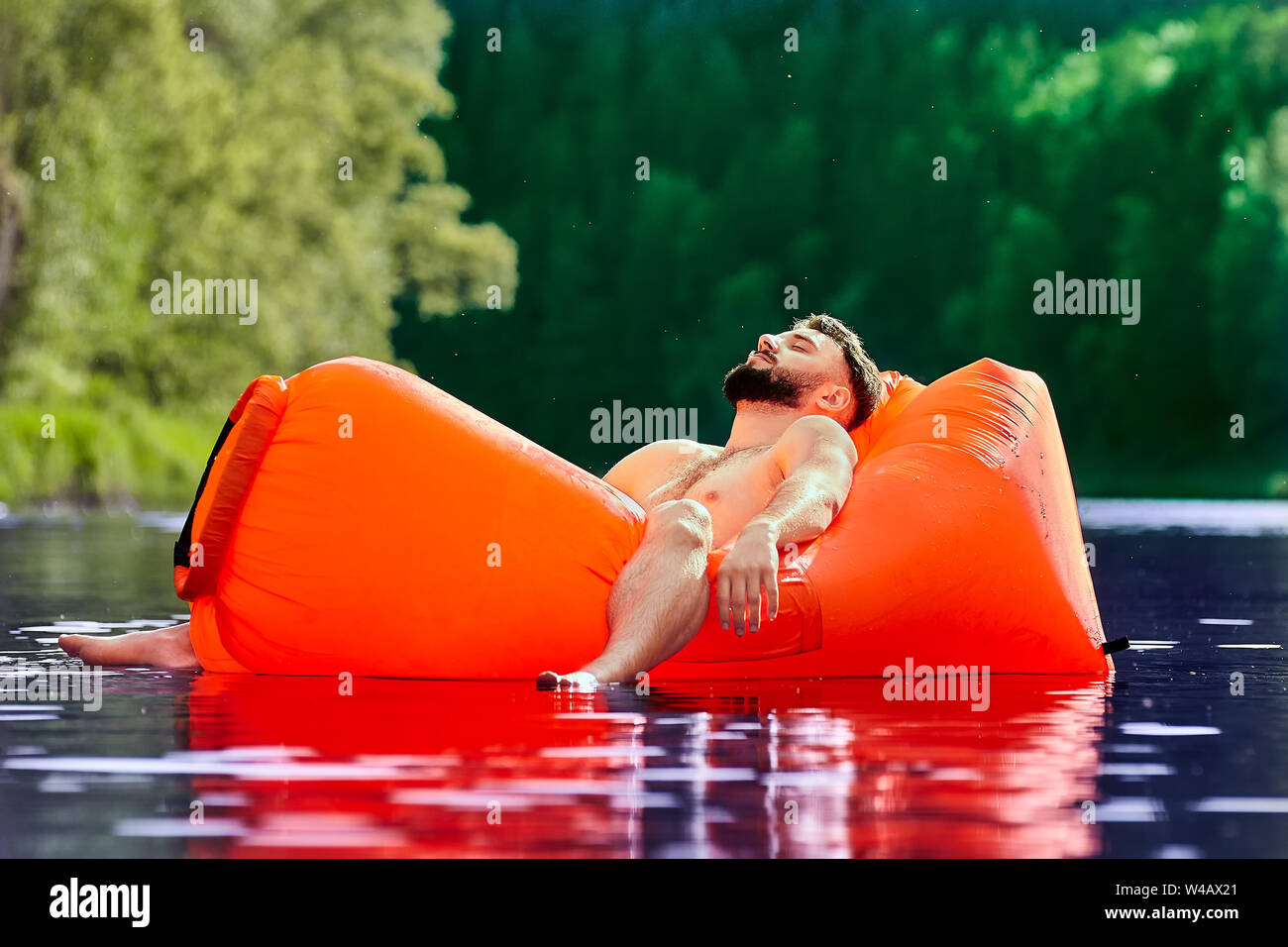 Un joven está descansando acostado en una tumbona naranja inflable flotando sobre la superficie del agua de un río del bosque. Camping durante el eco-turismo. Foto de stock