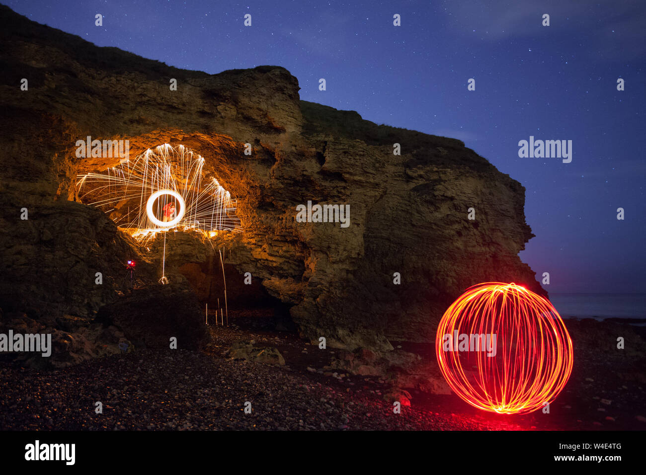 Dos amigos girando una rueda ardiente de fuego y un rojo brillante orbe durante una noche de aventura de la fotografía de la playa y las cuevas cercanas Foto de stock