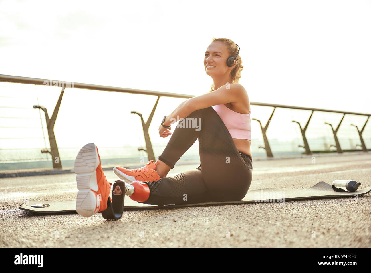 Feliz por la mañana. Atletas discapacitados positiva mujer con pierna prostética en auriculares haciendo ejercicios de yoga y sonriendo mientras estaba sentado en el puente. Deshabilitado el deporte concepto. La motivación. Estilo de vida saludable Foto de stock