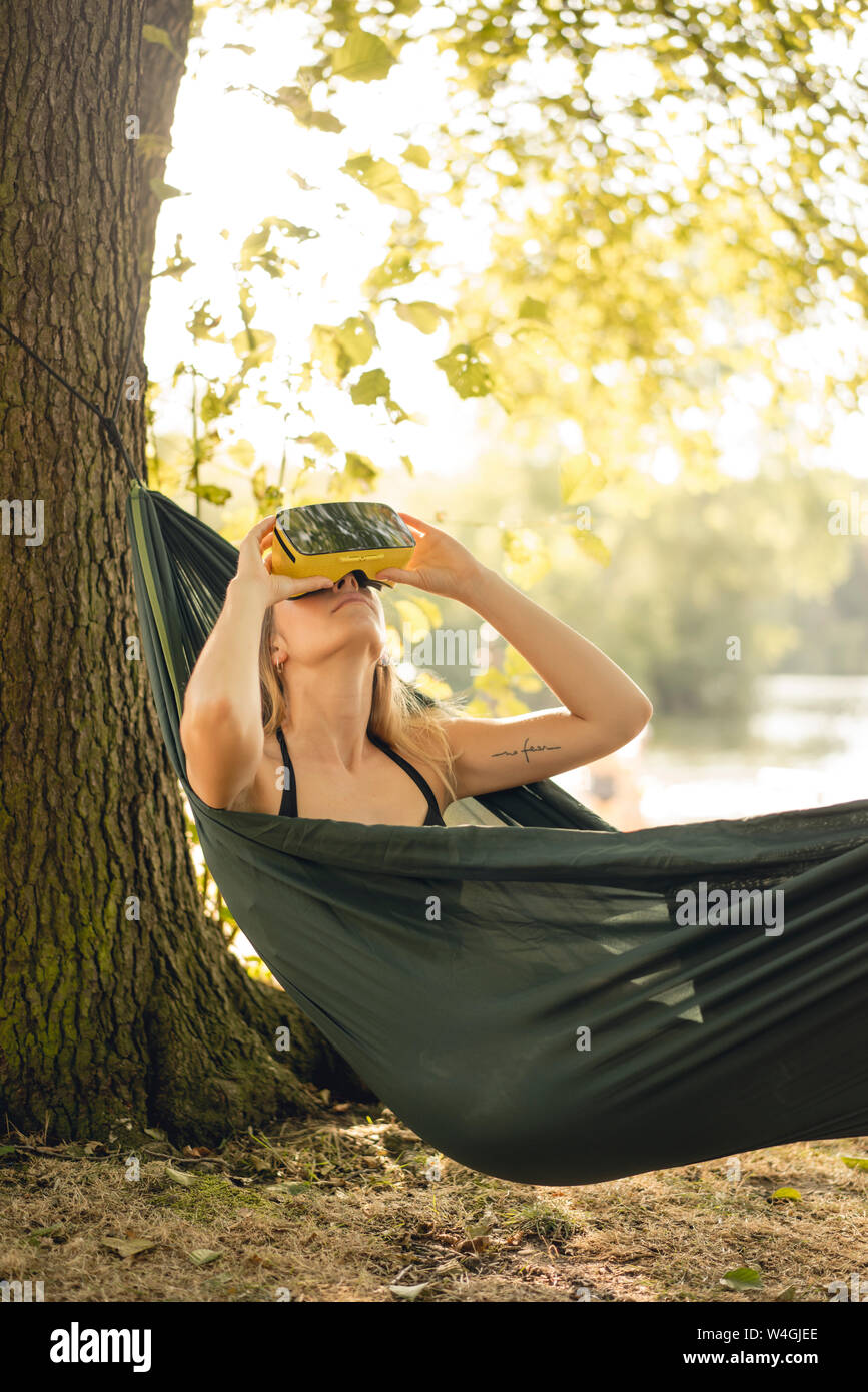 Mujer joven relajándose en una hamaca en el lago, utilizando gafas de VR Foto de stock