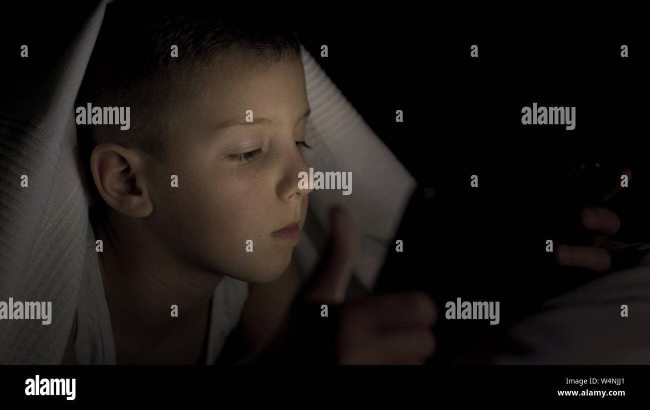Un muchacho feliz radica en la cama bajo una manta y juega en una tableta en un juego en la oscuridad. El rostro del niño está iluminado por un brillante monitor Foto de stock
