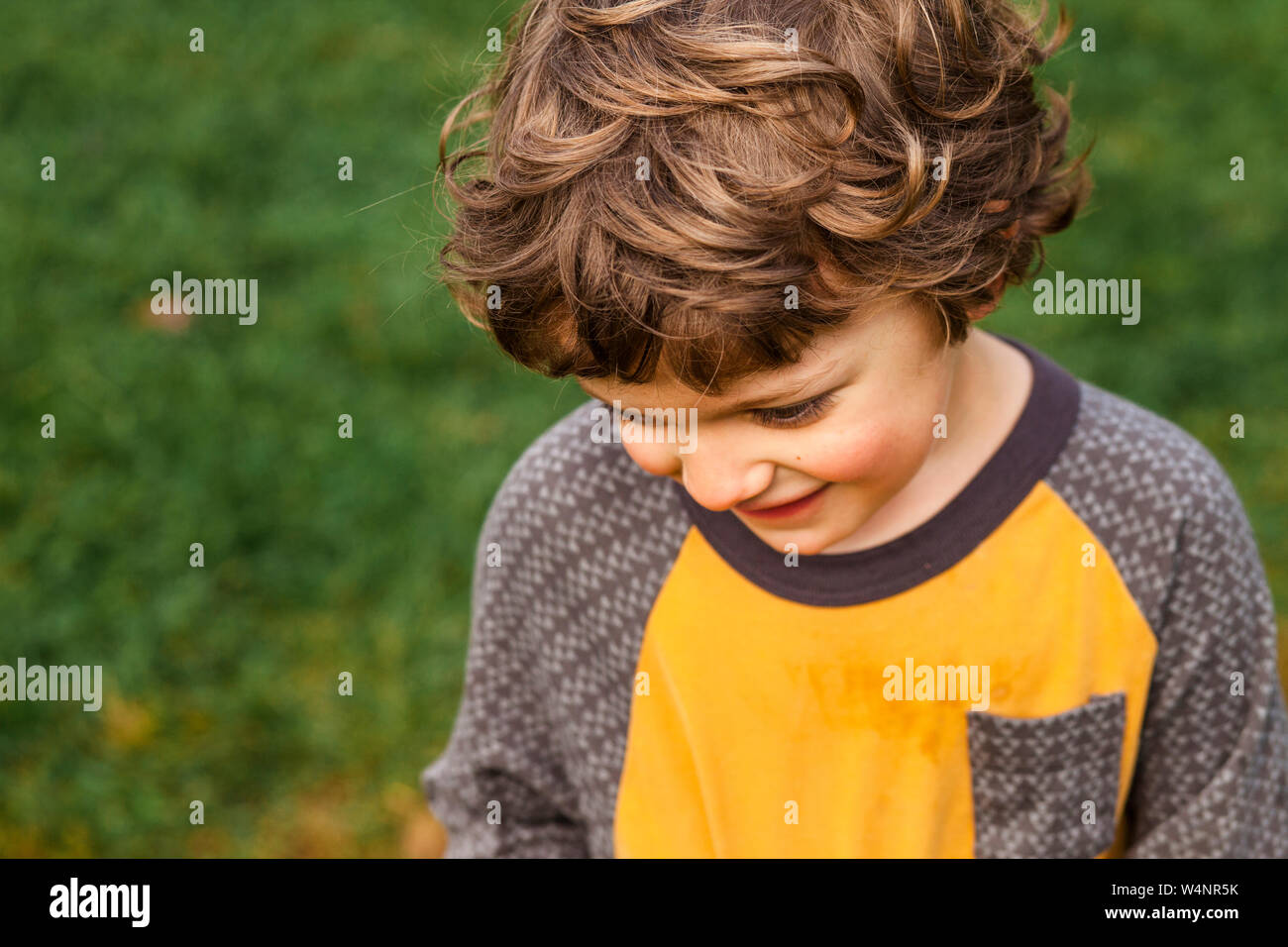 Un pequeño niño feliz con golden pelo rizado parece tímidamente hacia abajo Foto de stock