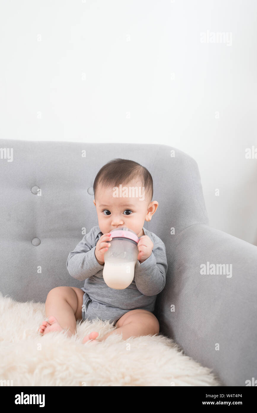Poco lindo bebé niña sentada en el sofá en la sala bebiendo leche de botella y sonriente. Bebé feliz. La gente de familia interior interior conceptos. B infantiles Foto de stock