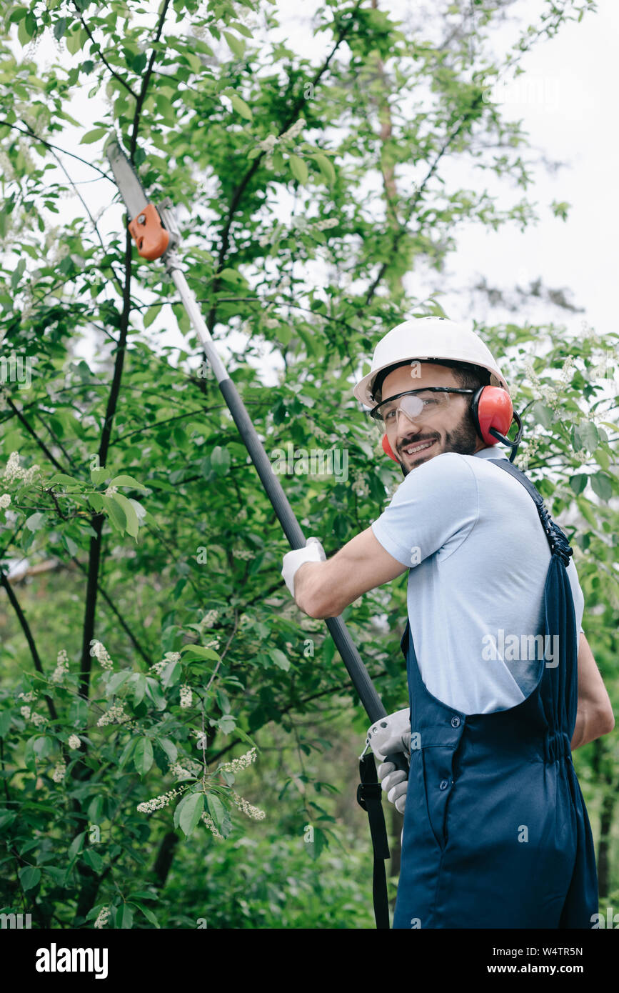 Sonriente jardinero en casco podar los árboles con pértiga telescópica vio  y mirando a la cámara Fotografía de stock - Alamy