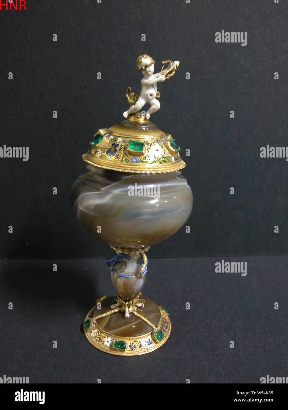 Cáliz y cubierta, cáliz, Cáliz con disparos, con los ojos vendados amor, Cáliz con tapa de onix, montado con esmaltados de oro y piedras preciosas. Una coronación, con los ojos vendados Amorphic figura de esmalte blanco como una corona. Amplia bordes dorados con una fina flor y pasillos de esmalte hundido con gemas superpuesta alrededor de la base y la tapa. El pie está conectado por tres tiras estrechas para el jarrón con forma de tallo, alrededor del cual a la altura del cuello, tres serpientes de viento, Cupido dispara un dardo, anónimos, Alemania, c. 1600 - c. 1620, Onyx (mineral), oro (metal), esmeralda (mineral), h 12,1 cm × 5,2 cm d d de 4,4 cm. Foto de stock