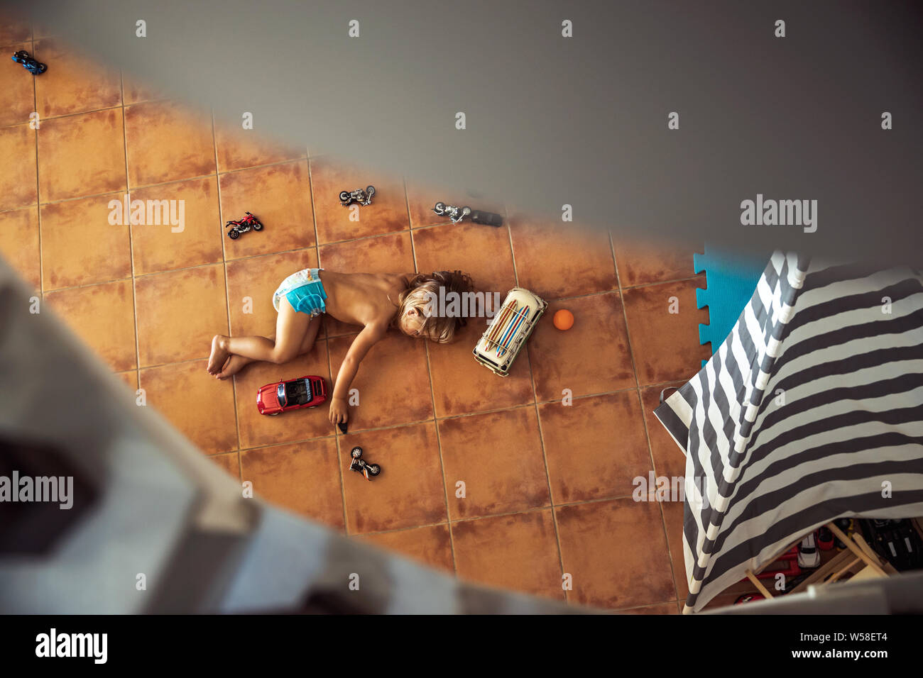 Chico se aburre en casa, tumbado en el suelo debajo de la escalera y jugar con modelos de autos, pasar sus vacaciones de verano en casa Foto de stock