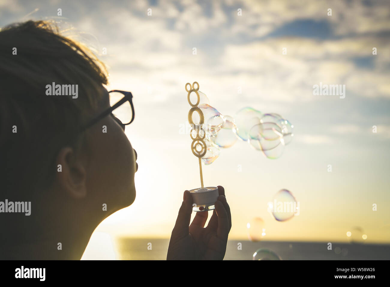 Muchacho joven rubia sopla una larga cadena de burbujas en un campo; lindo niño frente al océano persiguiendo jabón hierva, Niza kid jugar al aire libre en un hermoso Foto de stock