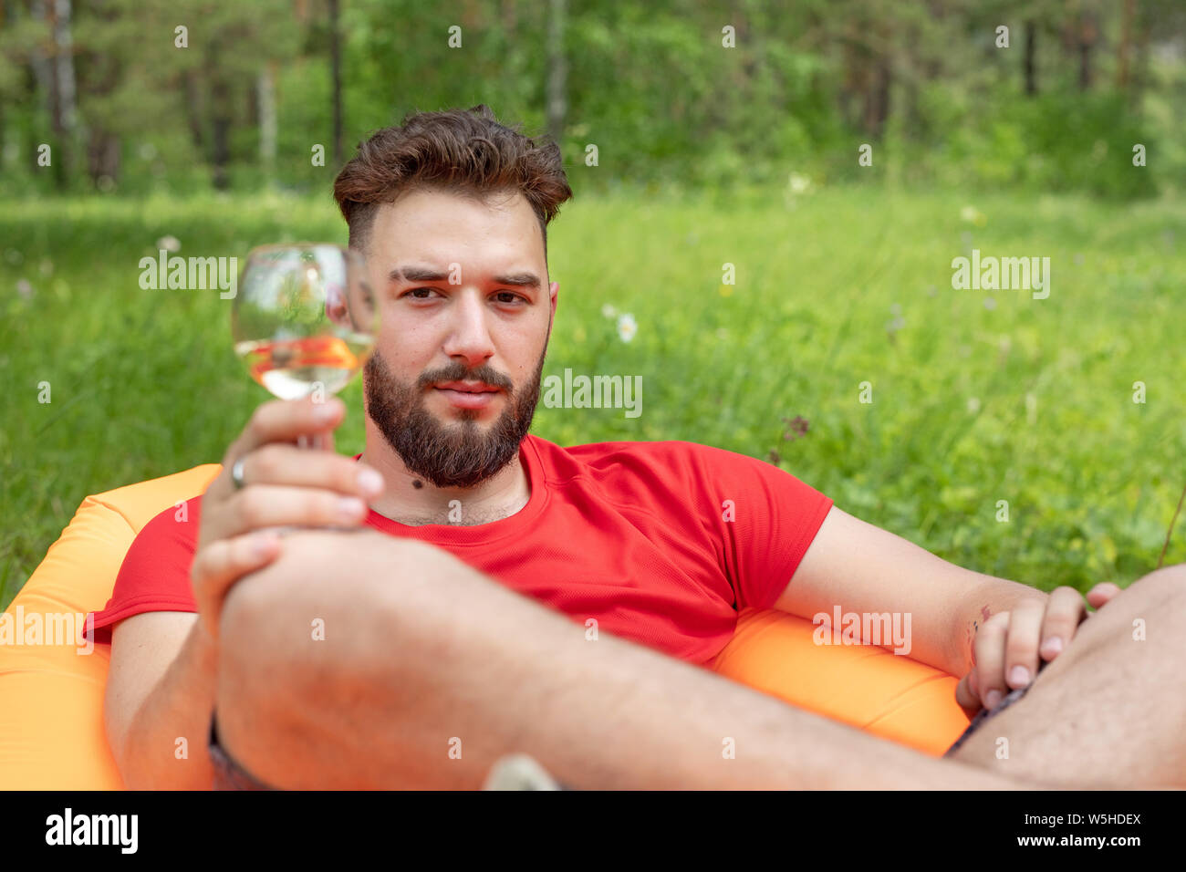 Concepto de ocio, camping, vacaciones de verano. Joven barbudo hombre caucásico con copas de vino sentando en el sofá naranja. Relajarse y disfrutar de la vida en el aire b Foto de stock