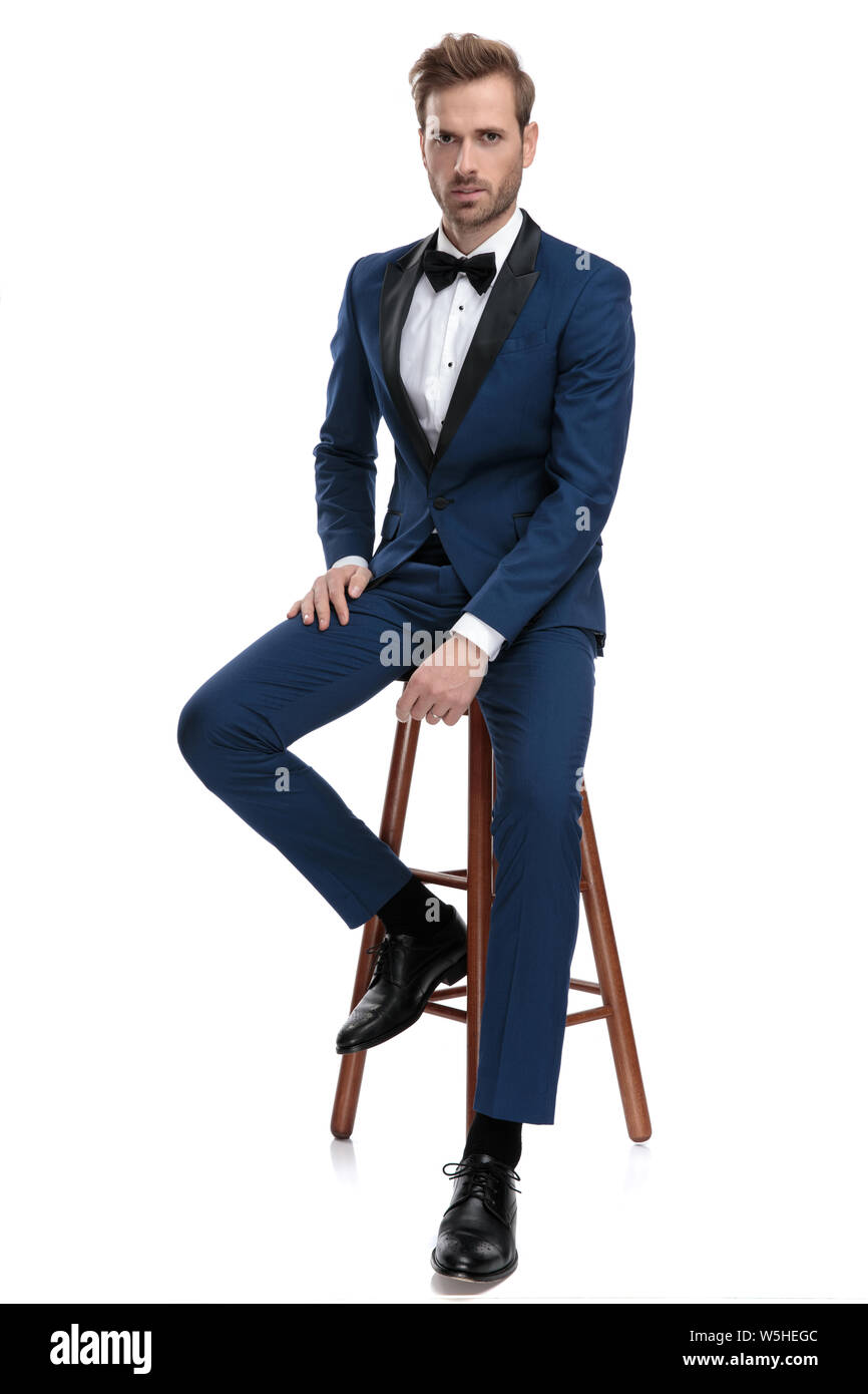 El hombre moderno en azul esmoquin sentado en un taburete con la mano en la  pierna sobre fondo blanco Fotografía de stock - Alamy