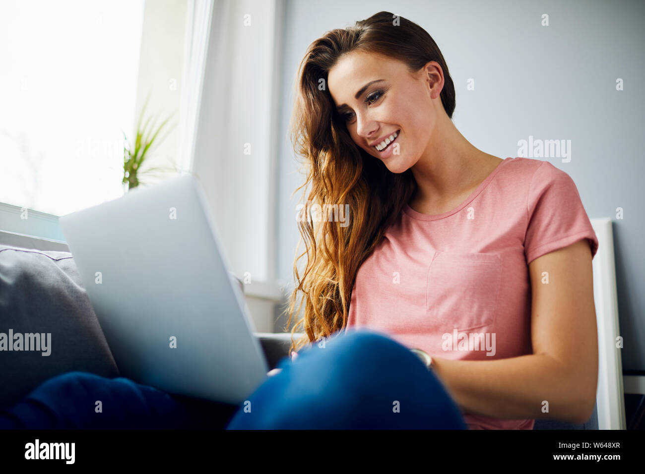 Retrato de una hermosa joven feliz y sonriente con el portátil mientras está sentado en el sofá en el salón Foto de stock