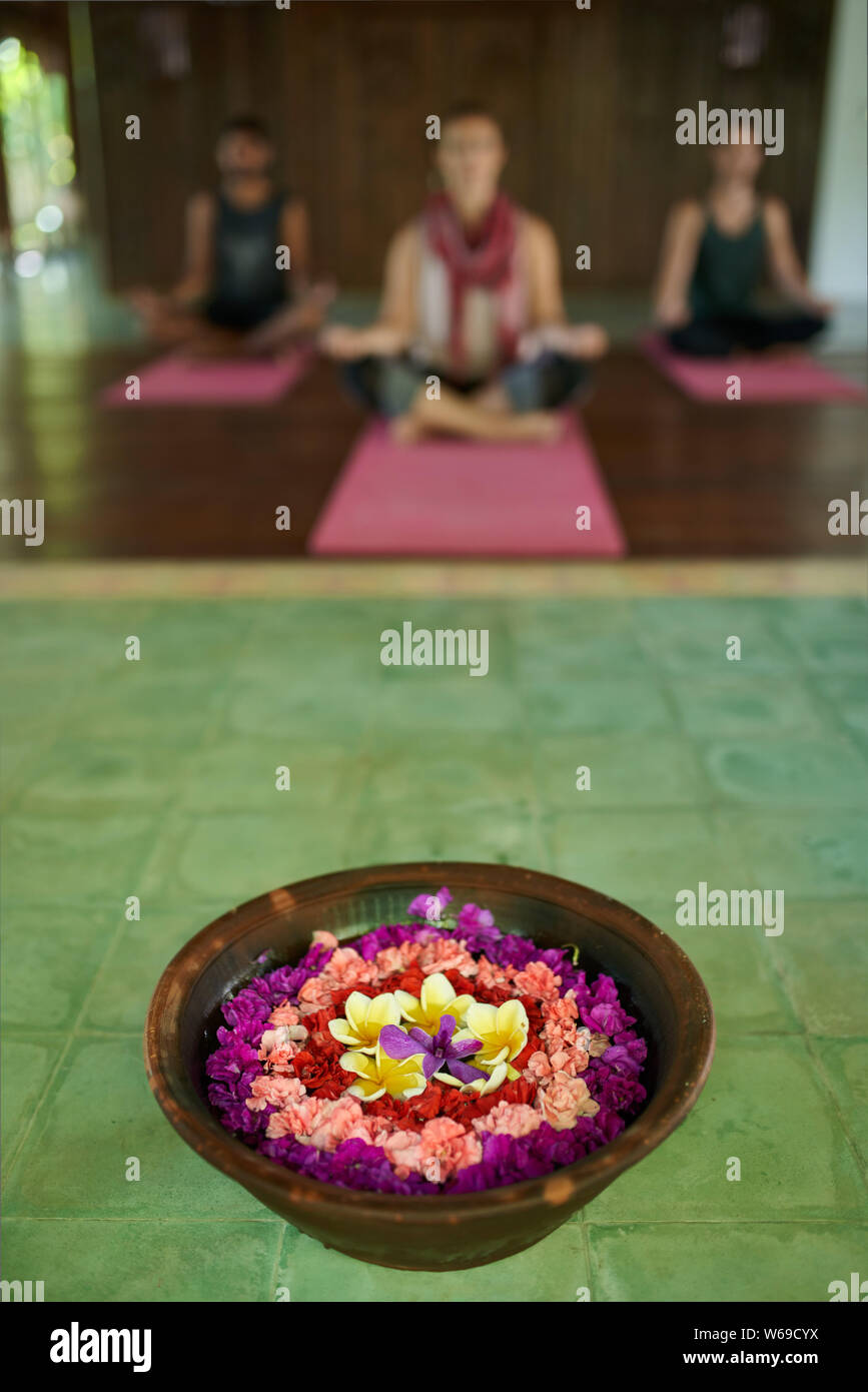 Cierre de la ofrenda de flores a la oración en el templo tradicional indonesio de Bali con tres diferentes personas meditando en segundo plano. Foto de stock