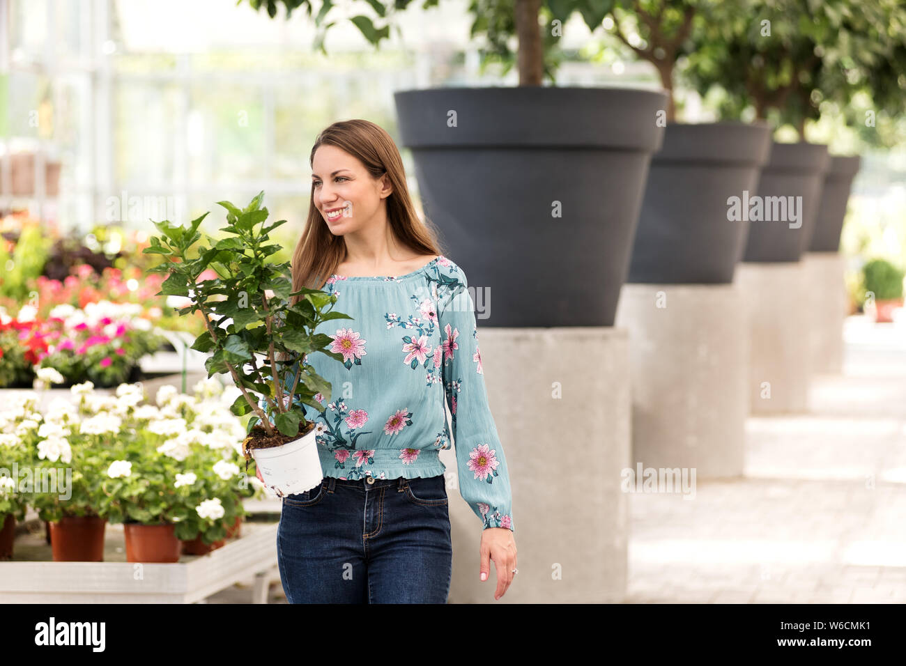 Atractiva mujer joven compra una planta Hibiscus en macetas en un vivero llevarla hacia abajo un pasillo con una sonrisa complacida, con espacio de copia Foto de stock