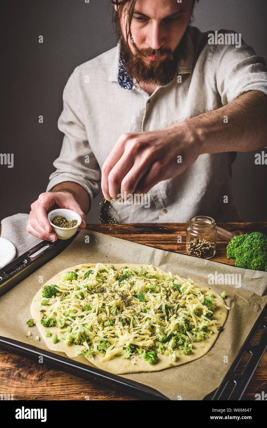 Hombre Barbado vierte especias sobre materias pizza con brócoli, salsa  pesto y queso Fotografía de stock - Alamy