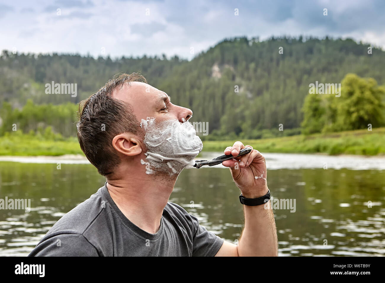 Hombre blanco con el rostro cubierto por espuma de afeitar es afeitarse por razor está en el bosque cerca del hermoso río, ecoturismo. Foto de stock