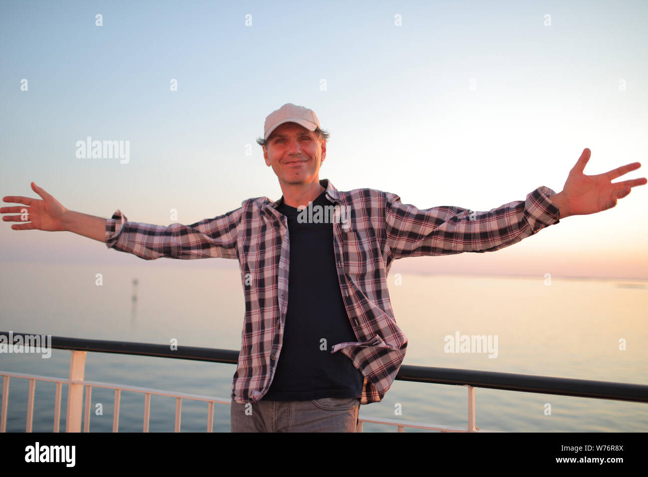 Feliz el hombre tiene los brazos extendidos en la cubierta del buque de crucero contra la puesta de sol sobre el mar tranquilo Foto de stock
