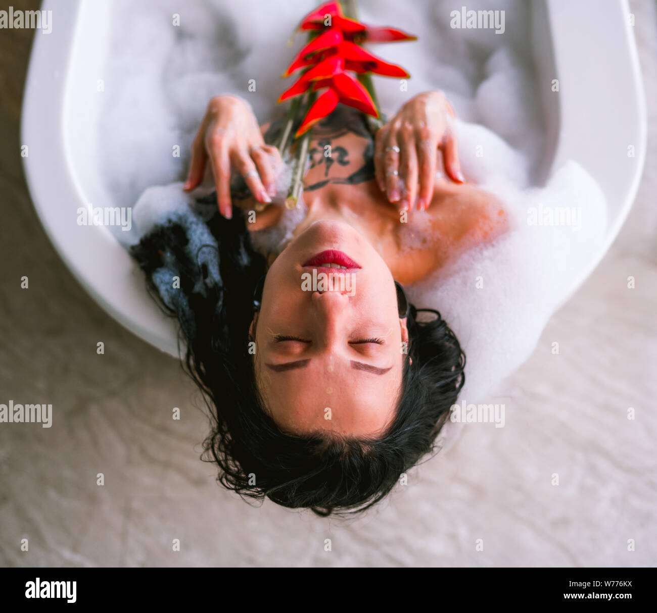 Mujer morena tomando el baño de burbujas en el baño luminoso con heliconia tropical flor roja Foto de stock