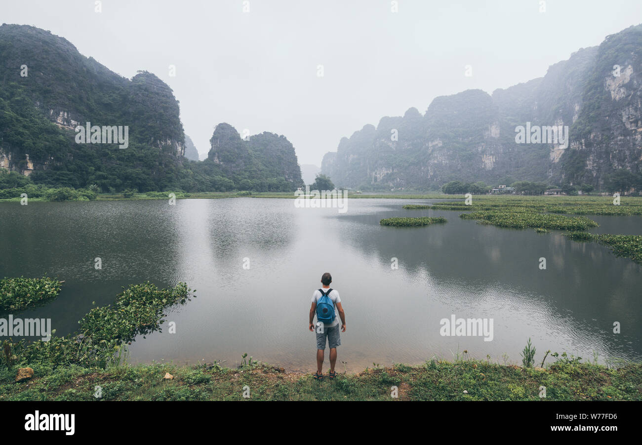 Hombre caucásico con vistas a las montañas de piedra caliza en la provincia de Ninh Binh, Vietnam. Día nublado, reflejo en el agua Foto de stock