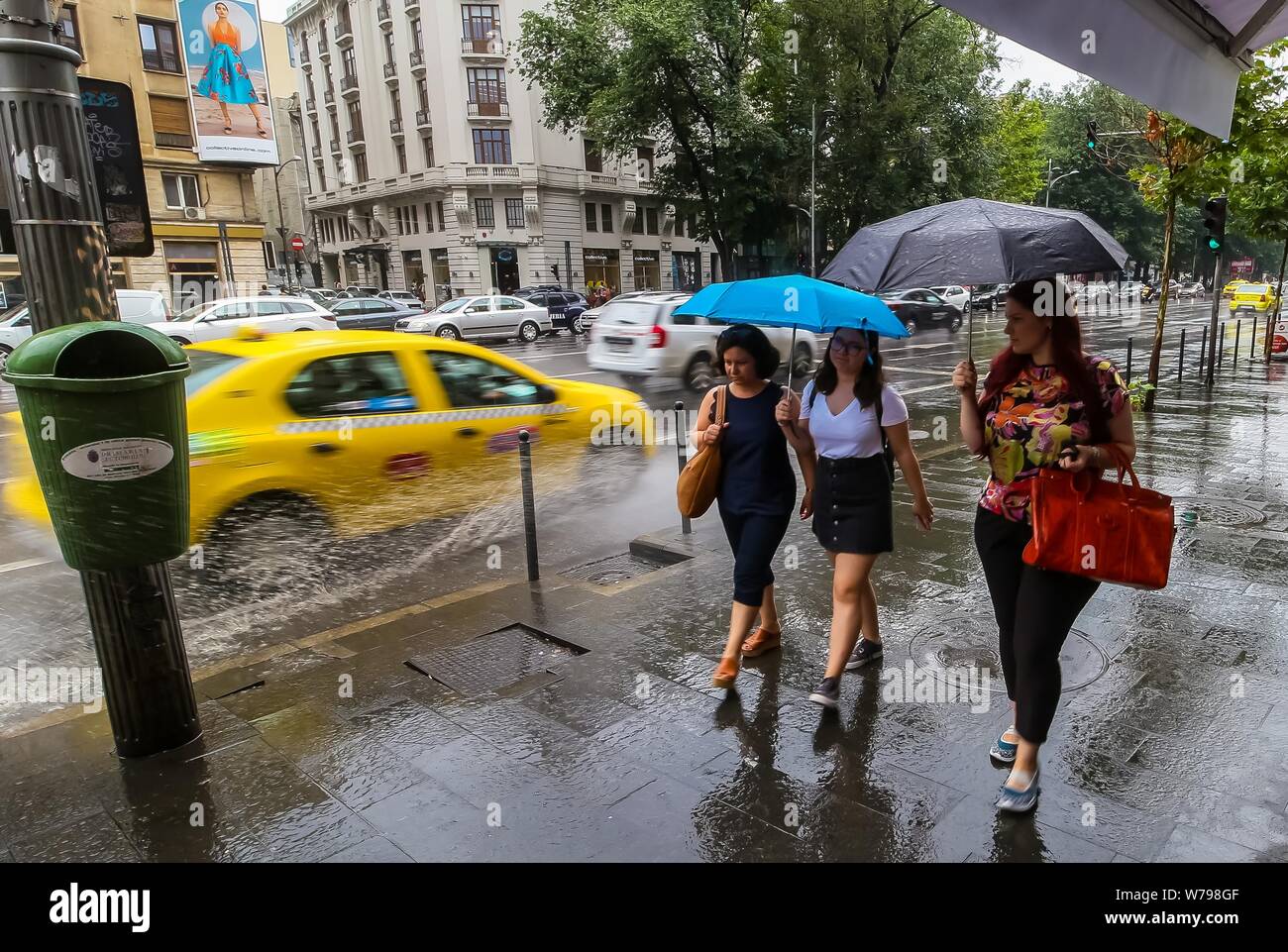 Bucarest, Rumania - 01 de agosto de 2019: Mujeres caminando con paraguas,  en tiempo lluvioso, mientras que un taxi las salpicó con agua de un charco  en el centro de la ciudad