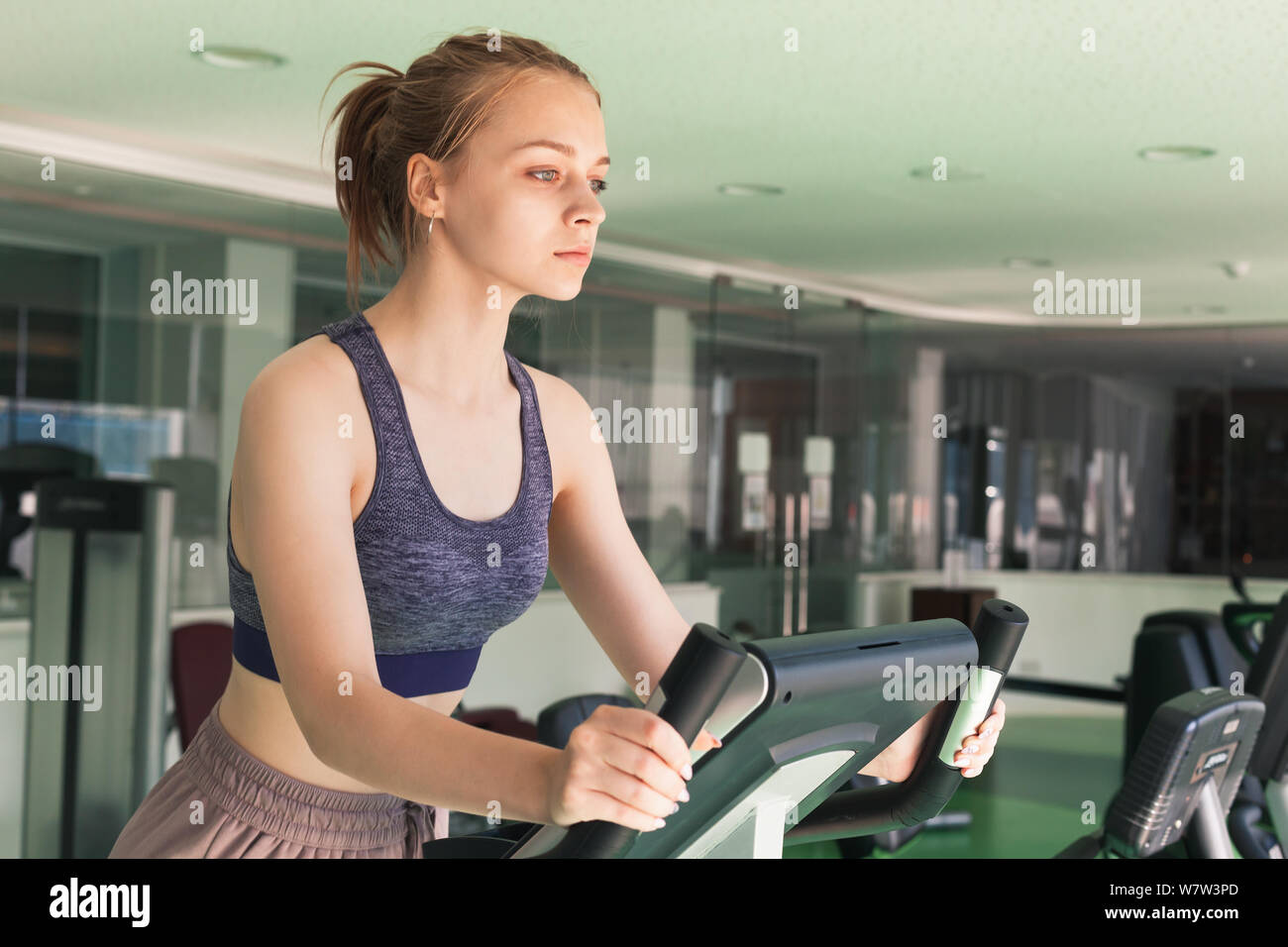 Joven rubia chica deportiva está metida en una bicicleta estacionaria en un gimnasio Foto de stock