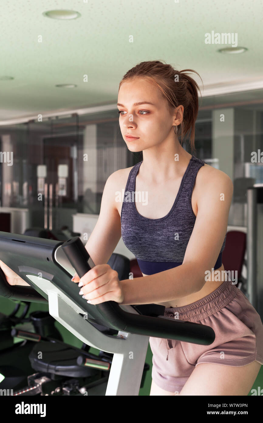 Hermosa joven rubia chica deportiva está metida en una bicicleta estacionaria en un gimnasio Foto de stock