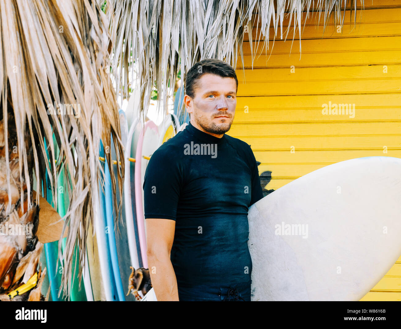 Retrato de un surfista en la cara de un protector solar, vistiendo pantalones cortos y una camiseta negra de pie con una tabla de surf. amarillo fondo de madera. Foto de stock