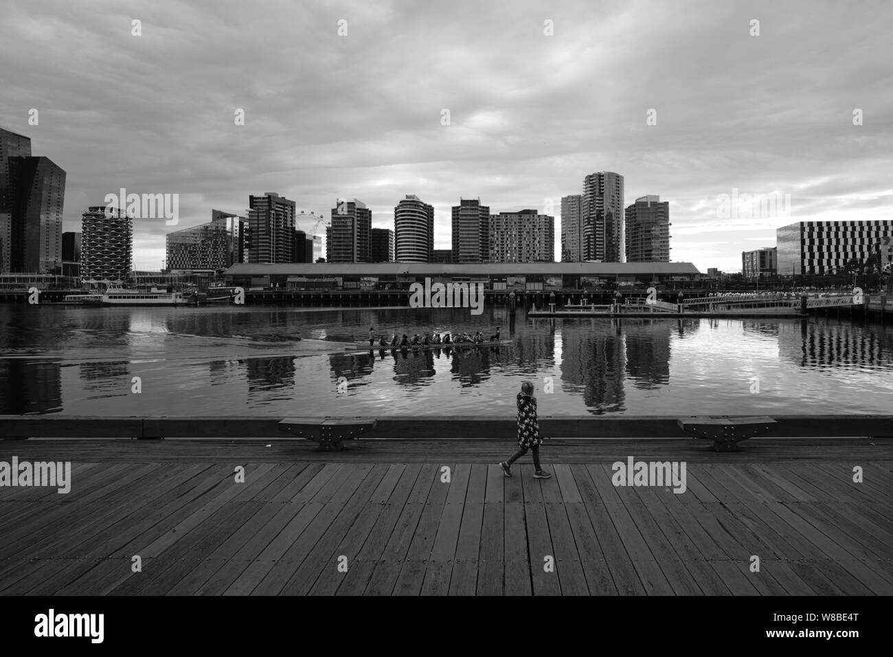 Adolescente caminando por el muelle en la zona de Docklands mientras ve remando palistas con edificios altos en el fondo. Melbourne, Australia Foto de stock