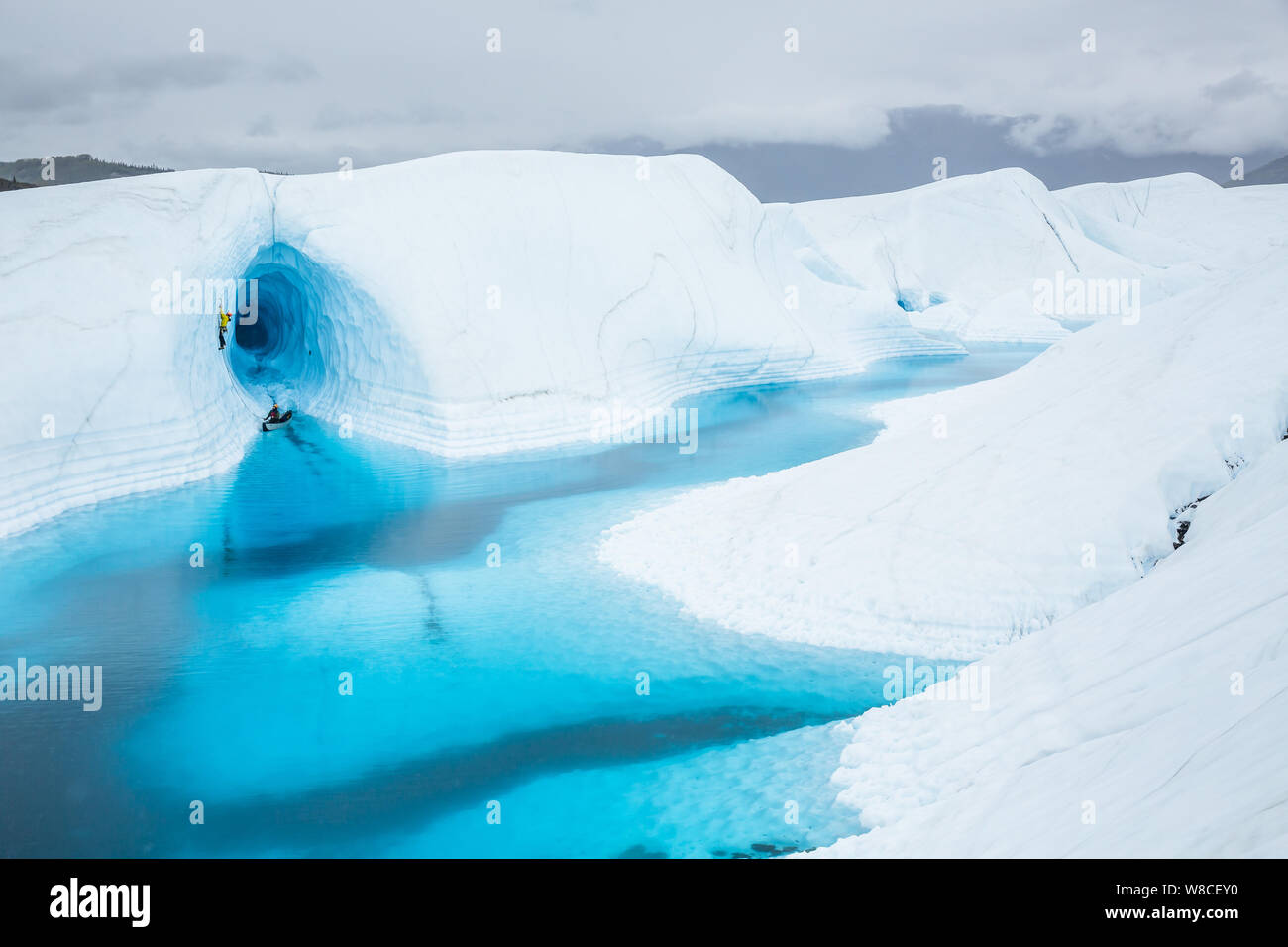 Escalador de hielo conduce a salir de una canoa delante de una gran cueva de hielo encima de una piscina azul. La piscina se encuentra en la parte superior del Glaciar Matanuska en Alaska. Foto de stock