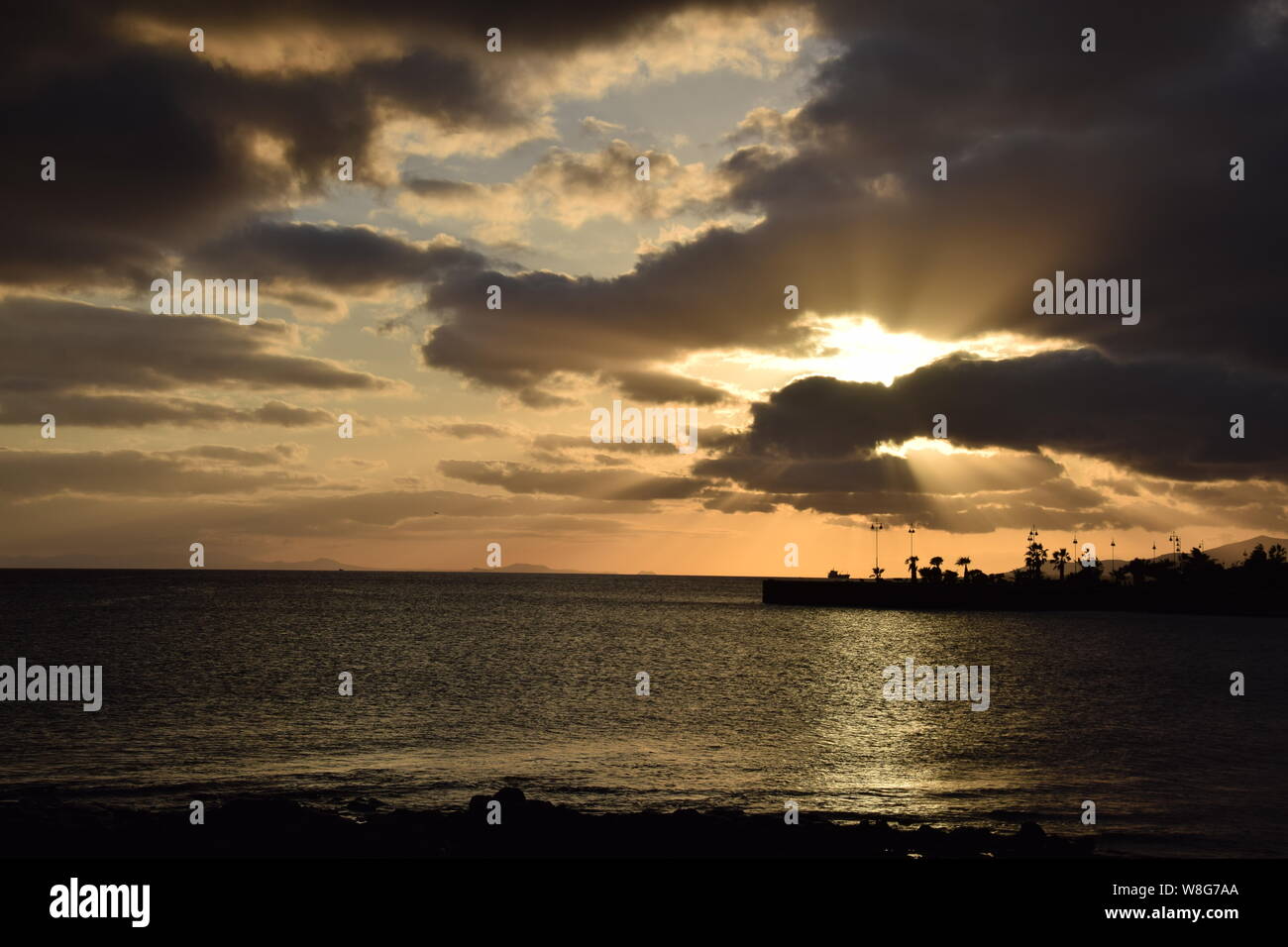 Rayos de sol a través de las nubes cerca de la puesta de sol / hora dorada, sobre el océano y el horizonte del parque frente al mar, Arrecife, Lanzarote Foto de stock