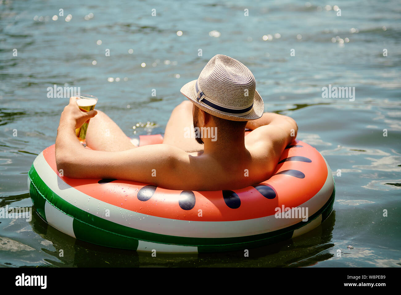 Los jóvenes caucásicos hombre con sombrero en la cabeza se encuentra a la deriva en el anillo inflable mientras se relaja en la naturaleza, hay una copa en su mano. Foto de stock