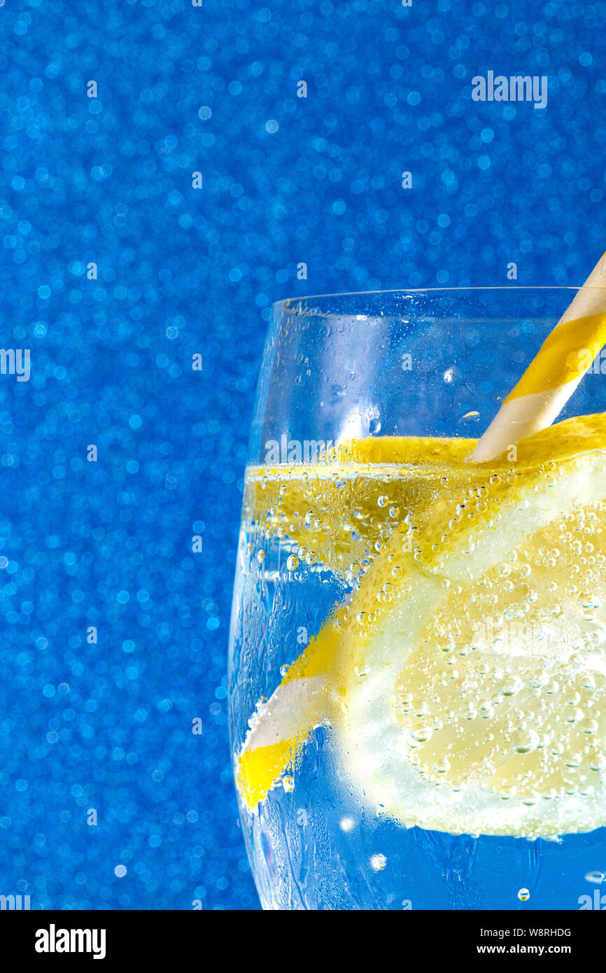 Soda fresca con burbujas un cubito de hielo, una rodaja de limón y una pajita en un fondo azul brillante Foto de stock